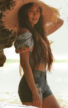 a woman wearing a straw hat standing on the beach with her arms behind her head