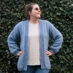 a woman standing in front of a hedge wearing a blue cardigan sweater and jeans