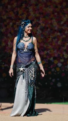 a woman with blue hair is dressed in an elaborate dress and headpiece, standing on the sand