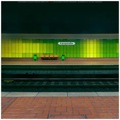 an empty train station platform with green and yellow lights on the wall, along with brick pavers