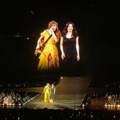 two women in yellow dresses standing on stage with one holding a guitar and the other singing