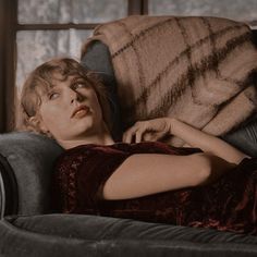 a woman laying on top of a gray couch next to a window with a blanket over her head