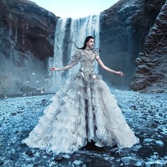 a woman standing in front of a waterfall wearing a long dress with feathers on it