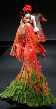 a woman in an orange and green dress walking down a runway with her hands on her hips