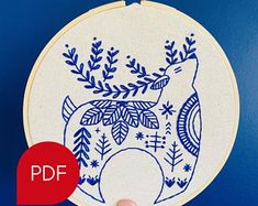 a hand is holding up a blue and white embroidery pattern on a hoop with an image of a deer