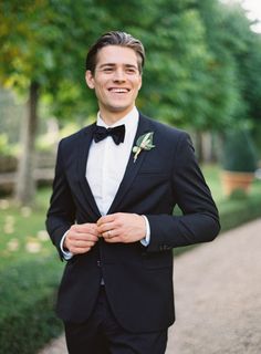 a man in a tuxedo smiles as he stands on a path near some trees