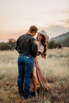 a man and woman standing in the middle of a field with tall grass at sunset