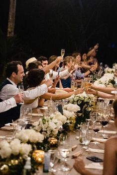 a group of people sitting at a long table with wine glasses in their hands and flowers on the table