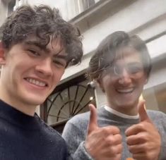 two young men standing next to each other giving the thumbs up sign in front of a building