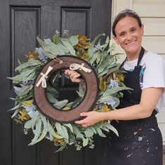 a woman holding a wreath with scissors on it in front of a black wooden door