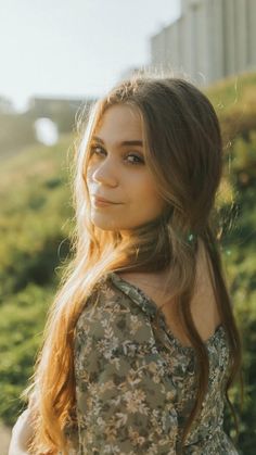 a young woman with long hair standing in front of some bushes and looking off into the distance