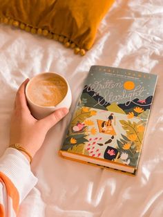 a person holding a cup of coffee next to a book on top of a bed