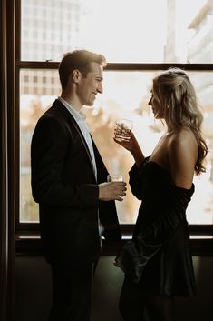 a man and woman standing next to each other in front of a window holding drinks