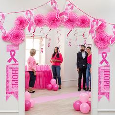 two women and one man are standing in front of a pink ribbon arch with balloons