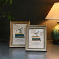 two framed pictures sitting on top of a table next to a green vase and lamp