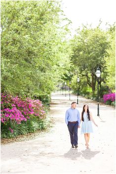 a man and woman walking down a path in the middle of some trees with pink flowers