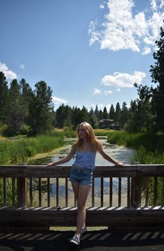 a woman is sitting on a bench looking at the water and trees in the background