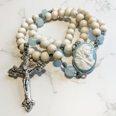 "This handcrafted, beautiful, and unique Catholic rosary is made with 8mm bleached white wood beads and 8mm aqua quartz beads. The beads are strung and knotted on micro cord (the slimmest type of paracord!) for durability. The cameo rosary center depicts the Madonna of the Streets. The crucifix is 2 inches in length and was made in Italy.  This rosary is about 14 inches in length and is lightweight and easy to carry with you so you can \"pray without ceasing.\" Each rosary from Refuge Rosaries is carefully packed in a cotton muslin drawstring bag that can be used as a rosary case. Follow us on Instagram and Facebook @refugerosaries to stay in touch and see our latest designs!" Rosary Case, Chaplet Rosary, Upcycled Vintage Jewelry, The Madonna, Catholic Rosary, Rosary Catholic, Catholic Gifts, Quartz Beads, Aquamarine Gemstone