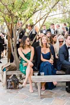 two women sitting on a wooden bench in front of a group of people wearing tuxedos