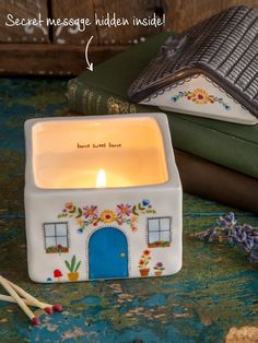 a small white candle holder with a house on it next to some books and candles