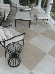 an outdoor seating area with white cushions and black iron furniture on tiled flooring, surrounded by greenery