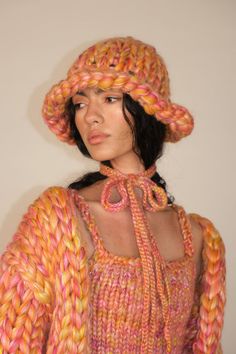 a woman wearing a knitted hat and dress with braids on the sides, standing in front of a white wall