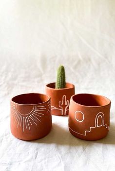 three clay pots with designs on them and a cactus in the middle, sitting on a white sheet