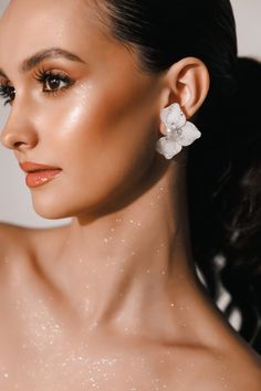 a woman wearing white flower earrings on her left ear, with glittery skin and dark hair