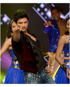 a man in a black shirt and blue skirt pointing at the camera with other dancers behind him