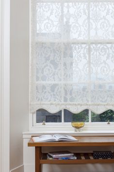 a table with books and a vase on it in front of a window that has lace curtains