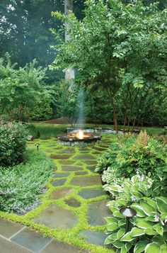 an outdoor fire pit surrounded by lush green trees and shrubs in the background is a stone path that leads to a well manicured garden