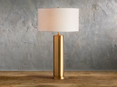 a gold lamp on a wooden table with a gray wall behind it and a white shade