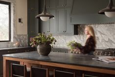 a woman standing in a kitchen next to a large potted plant on top of a counter