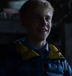 a young man with blonde hair wearing a blue and yellow jacket smiling at the camera