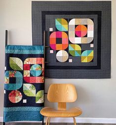 two quilts are hanging on the wall next to a chair and a framed art piece