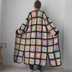 a woman in a crocheted coat is standing near a wall and looking at the floor