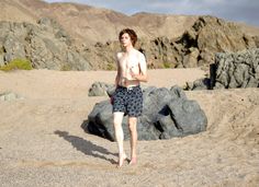 a shirtless man walking on the beach with rocks in the foreground and mountains in the background