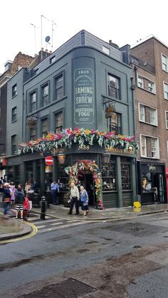 people walking on the sidewalk in front of a building with flowers hanging from it's roof