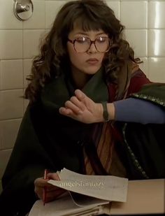 a woman with glasses sitting at a table in front of a white tile wall and writing