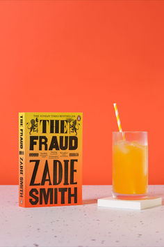 an orange juice in a glass next to a copy of the fraud by zadie smith