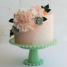 a pink cake with flowers on top sitting on a green pedestal in front of a white brick wall