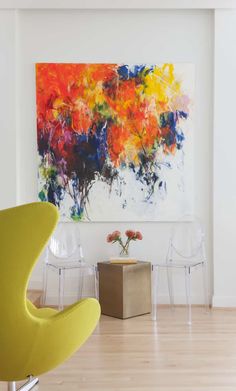 an abstract painting hangs on the wall next to two chairs and a vase with flowers in it