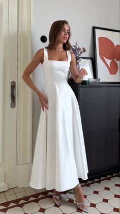 a woman in a white dress standing next to a cabinet with flowers on top of it