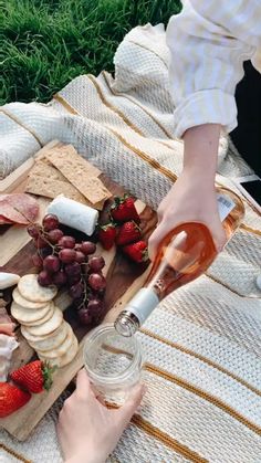 a person pouring wine into a glass on top of a cutting board filled with cheese, fruit and crackers