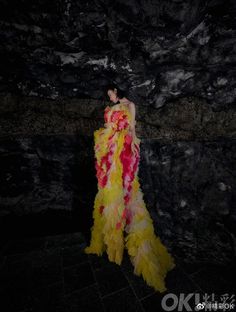 a woman in a long yellow and pink dress standing next to a rock wall at night