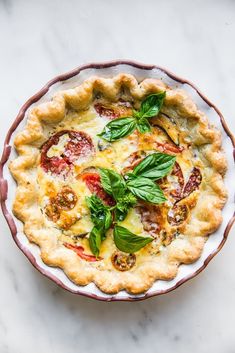 a pie with cheese, tomatoes and basil on top in a white dish sitting on a marble surface