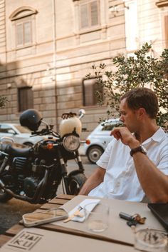 a man sitting at an outdoor table in front of a parked motorbike and motorcycle