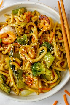 a white bowl filled with noodles, shrimp and veggies next to chopsticks