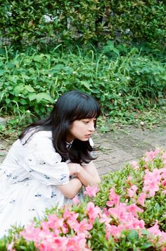 a woman sitting on the ground in front of some bushes and flowers with her head down