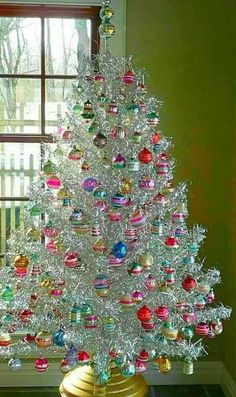 a white christmas tree sitting in front of a window next to a gold vase filled with ornaments